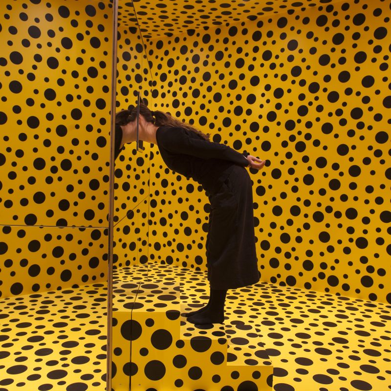 Foto de instalação de Yayoi Kusama usada como ilustração da ideia de "Lembrar de Quase Nada". Um quarto todo pintado de bolinhas com uma mulher ao centro olhando dentro de uma caixa espelhada.